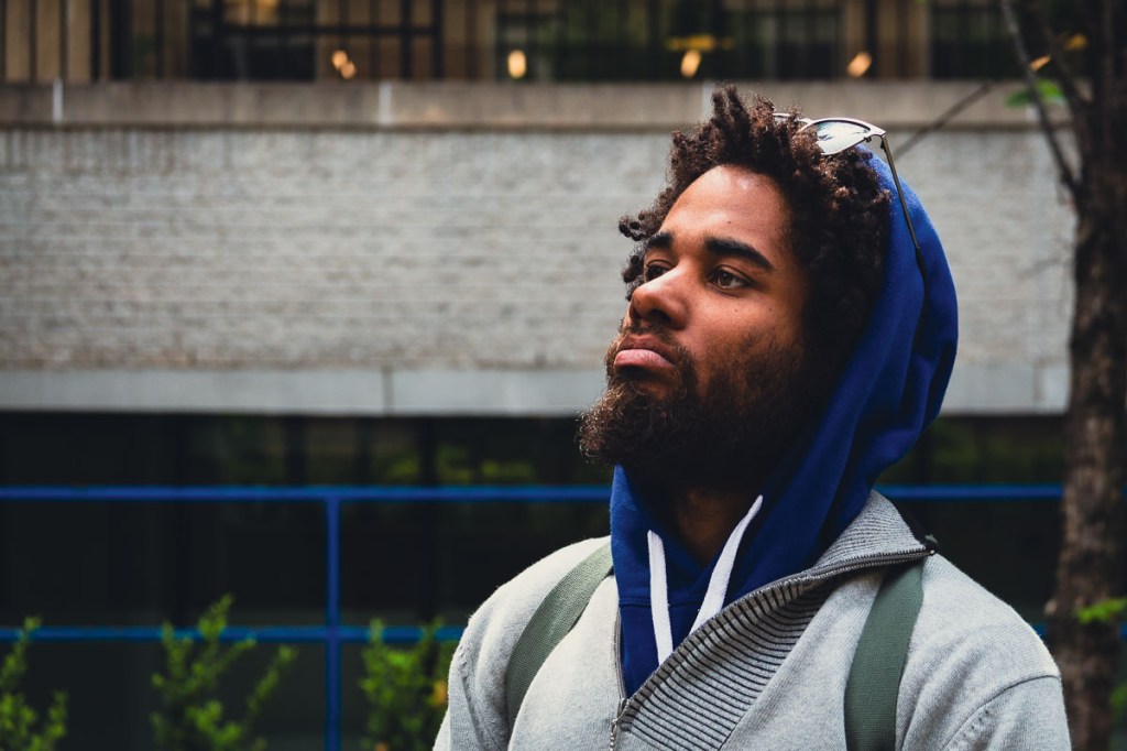 Black male wearing a blue hoodie under a grey sweater and a backpack