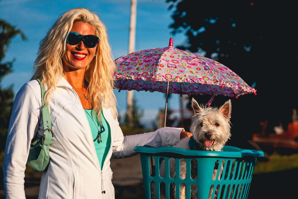 Platinum blonde woman wearing sunglasses with her Terrier in a teal basket and pink patterned umbrella