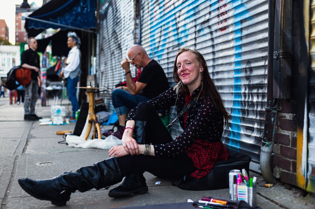 Female artist smiling and sitting comfortably on sidewalk with colourful markers in front and graffiti behind. © Andy Lee, 2022. realandylee.com