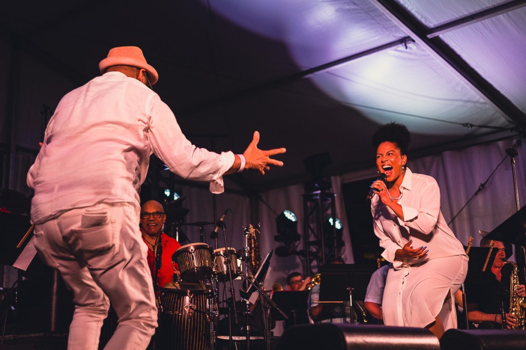 A Black woman in a white shirt dress crouches while singing to a man dressed in white. © Andy Lee, 2022. realandylee.com