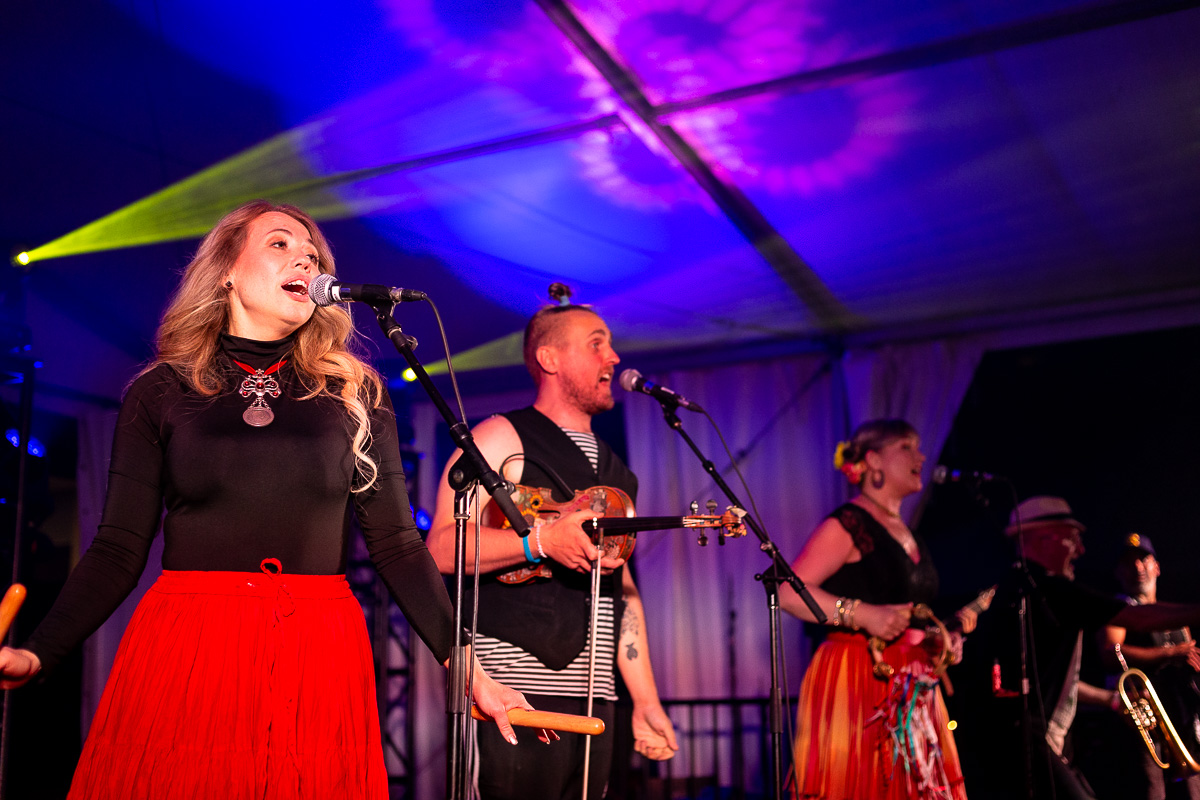 A blonde woman, a man with a violin and fellow bandmates sing inside a festival tent. © Andy Lee, 2022. realandylee.com