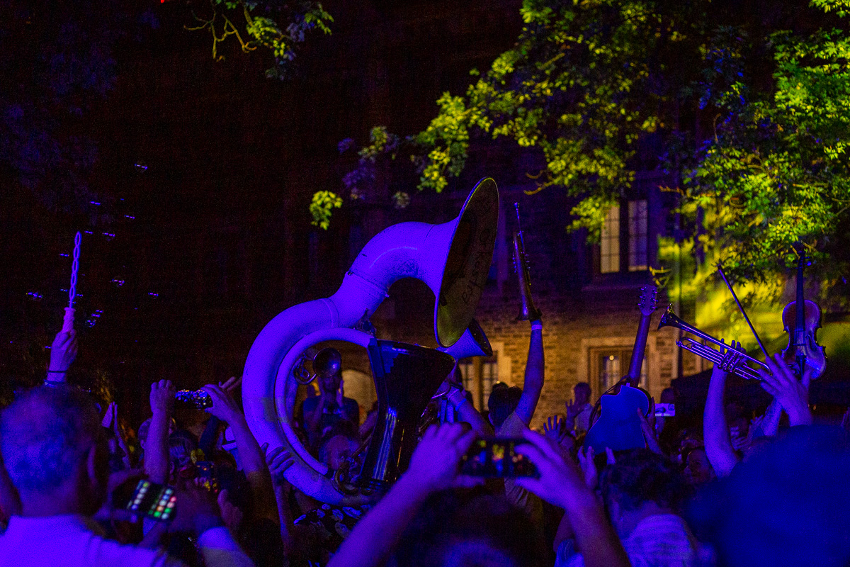 A band holds up a sousaphone, hand drum, trumpets, guitar and violin in the middle of a crowd with trees above at night. © Andy Lee, 2022. realandylee.com