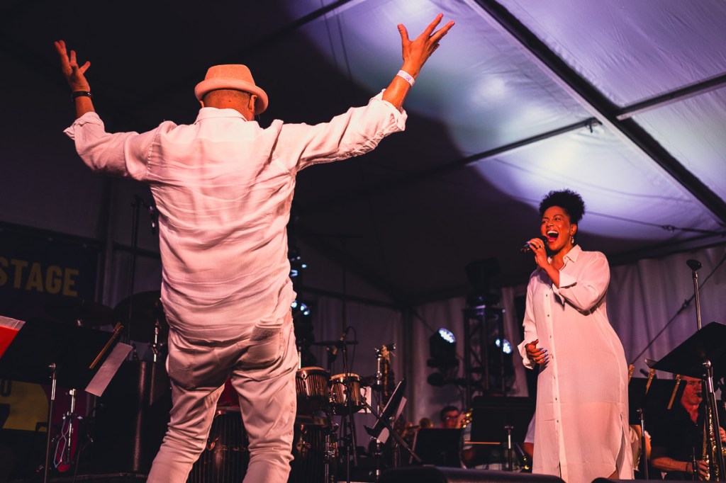 A Black woman in a white shirt dress sings to a man dressed in white raising his hands above his head. © Andy Lee, 2022. realandylee.com