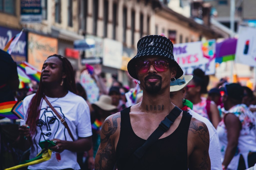 A tattooed Black man wearing pink sunglasses walks during the Dyke March in Toronto. © Andy Lee, 2022. realandylee.com