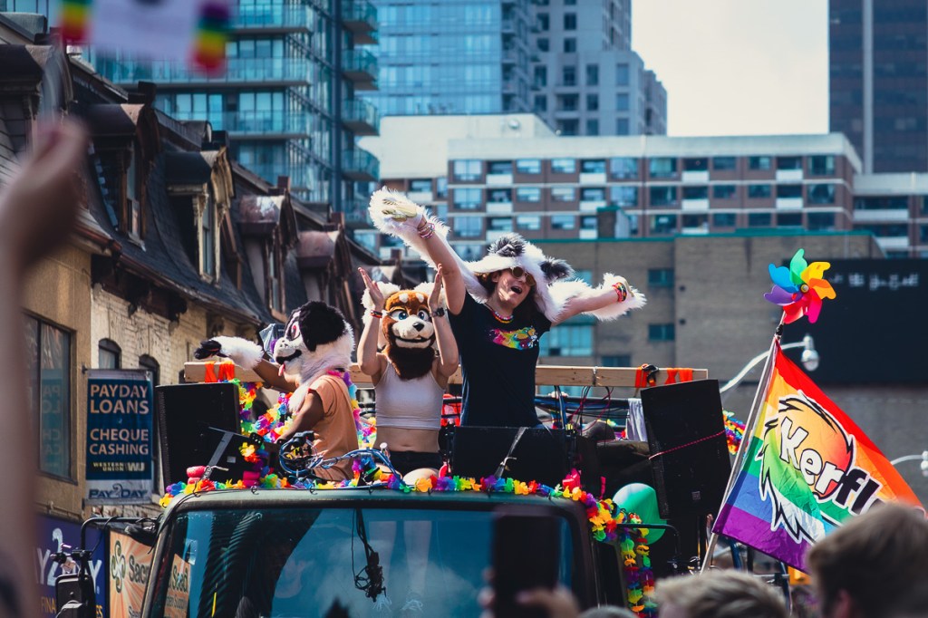 Two white women and a Black man wearing furry costumes atop a float at the Toronto Pride Parade. Onlookers watch the Toronto Pride Parade from their second-storey apartment windows, decorated with a rainbow flag. © Andy Lee, 2022. realandylee.com