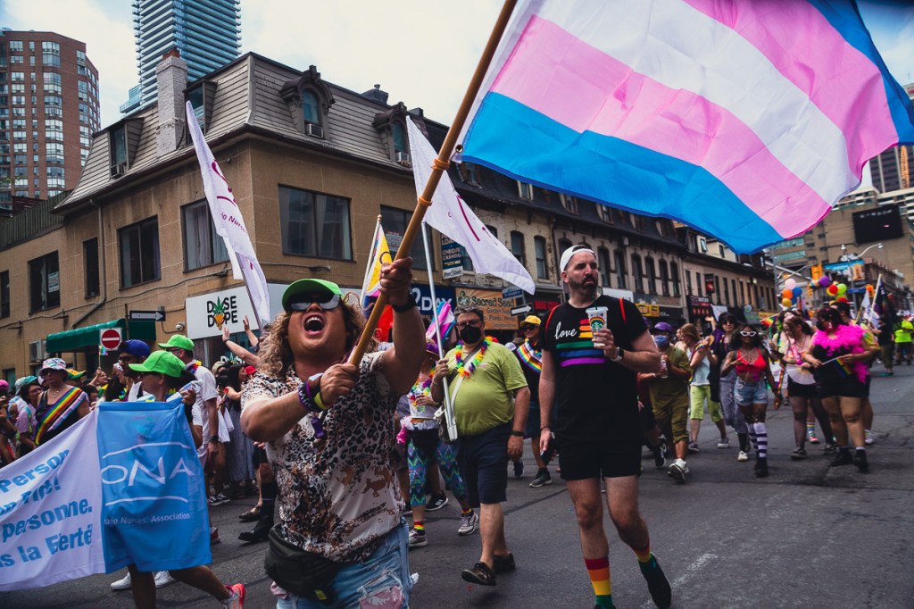 A man waves a large blue, pink and white flag at the Toronto Pride Parade. Onlookers watch the Toronto Pride Parade from their second-storey apartment windows, decorated with a rainbow flag. © Andy Lee, 2022. realandylee.com