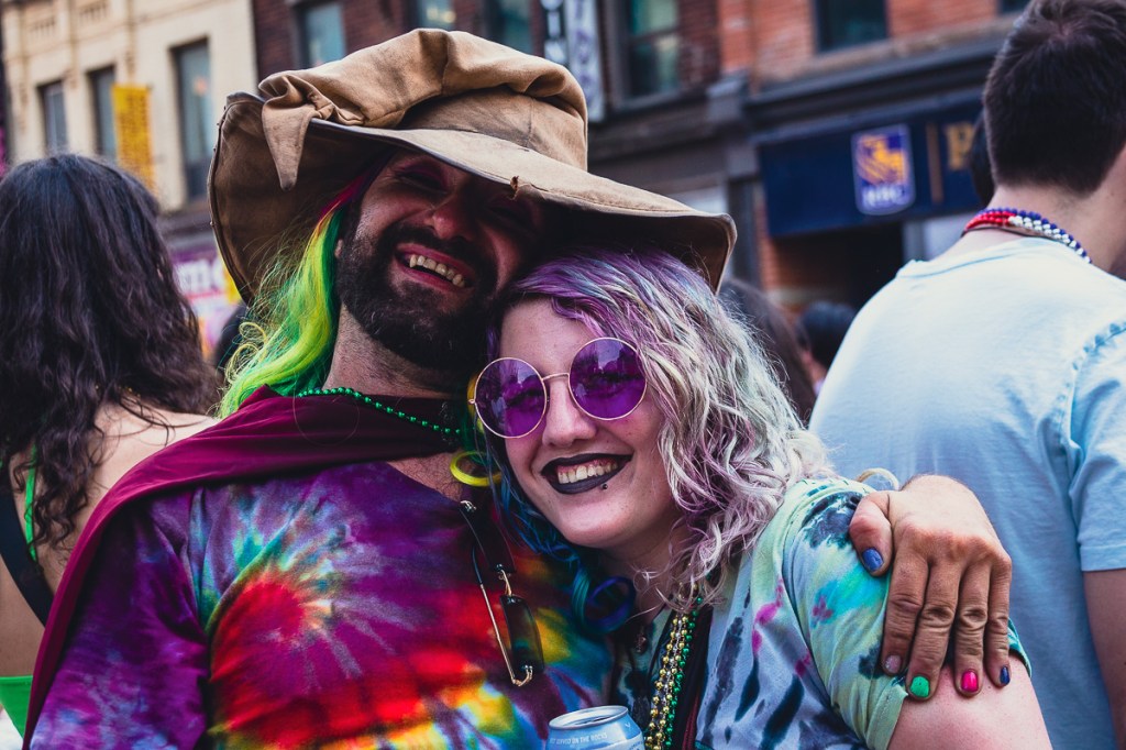A couple with colourful dyed hair and tie-dyed T-shirts at the Toronto Pride Parade. Onlookers watch the Toronto Pride Parade from their second-storey apartment windows, decorated with a rainbow flag. © Andy Lee, 2022. realandylee.com