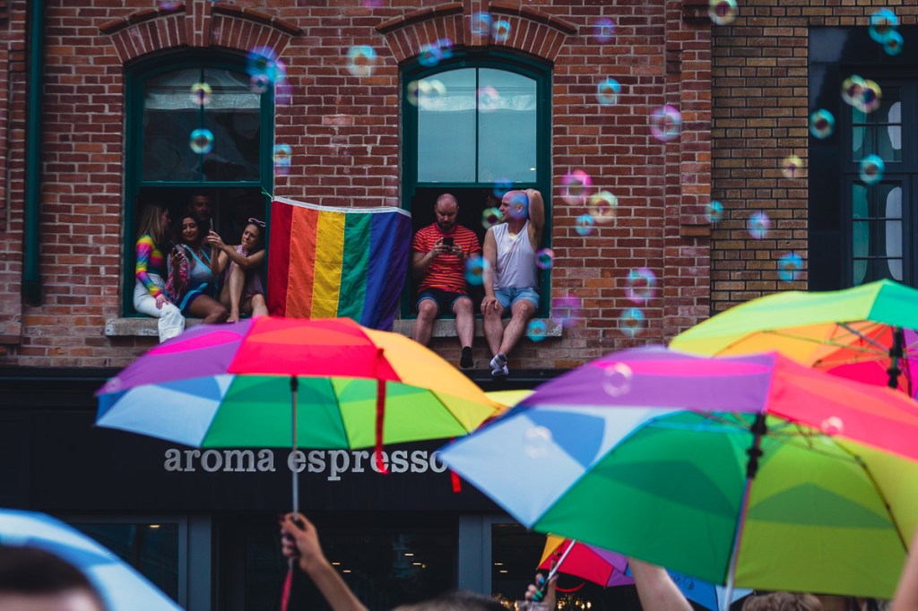 Onlookers watch the Toronto Pride Parade from their second-storey apartment window, decorated with a rainbow flag, as rainbow-coloured umbrellas and bubbles float by. © Andy Lee, 2022. realandylee.com