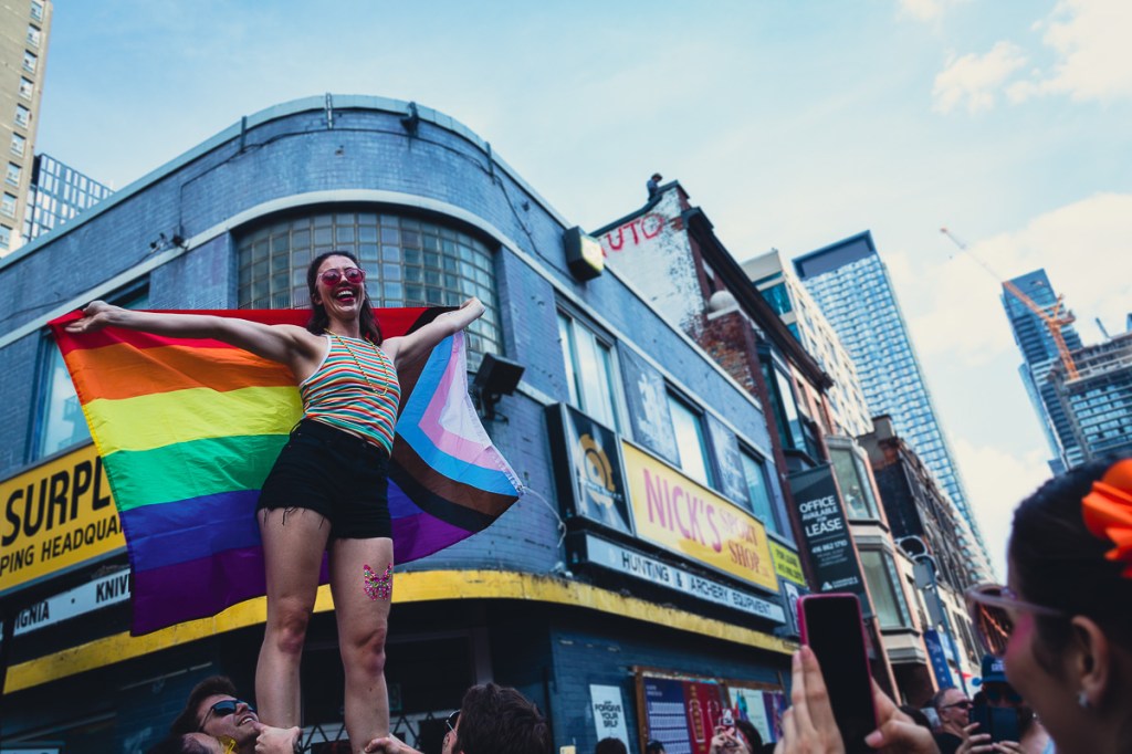 A smiling woman wearing pink sunglasses and a multi-coloured striped tank top holds a rainbow flag behind while her friends lift her above a crowd on a downtown street. © Andy Lee, 2022. realandylee.com