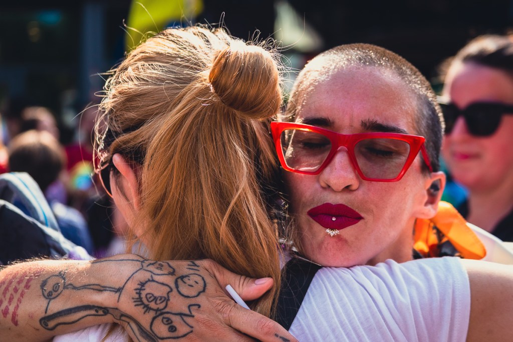 A woman with a shaved head, red sunglasses, labrette piercing and tattooed right forearm hugs a blonde woman during the Toronto Pride parade. © Andy Lee, 2022. realandylee.com