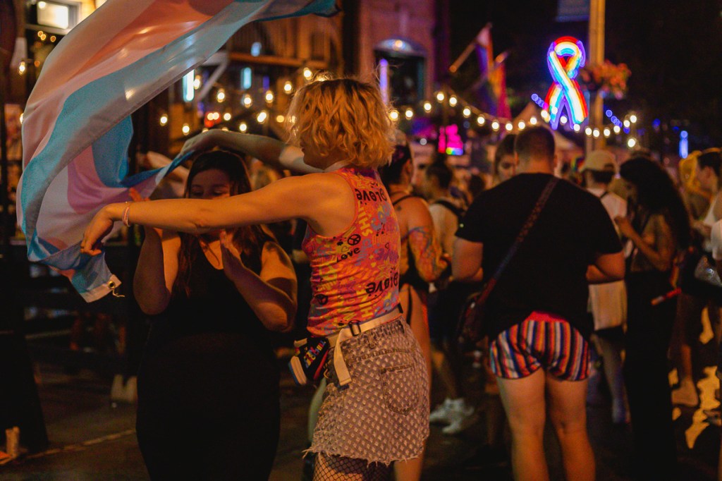A blonde woman waves a blue, pink and white flag over a brunette woman on the street at night. © Andy Lee, 2022. realandylee.com