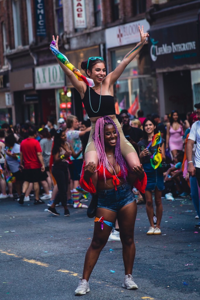 A white woman holds up peace signs while sitting on a Black woman's shoulders at the Toronto Pride Parade. © Andy Lee, 2022. realandylee.com