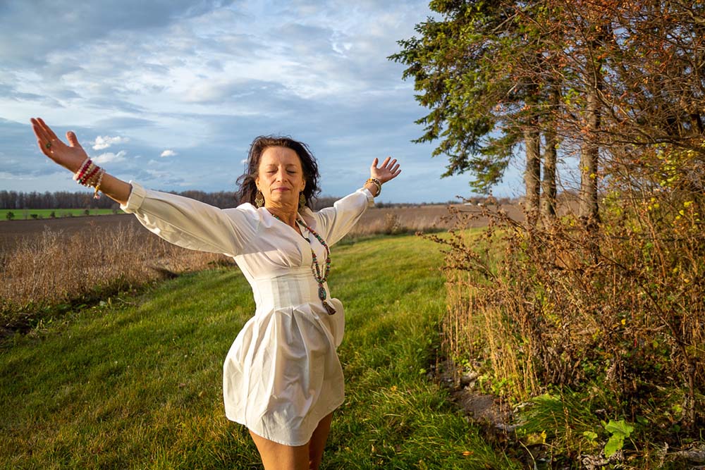 Brunette woman wearing a white dress with arms raised in nature