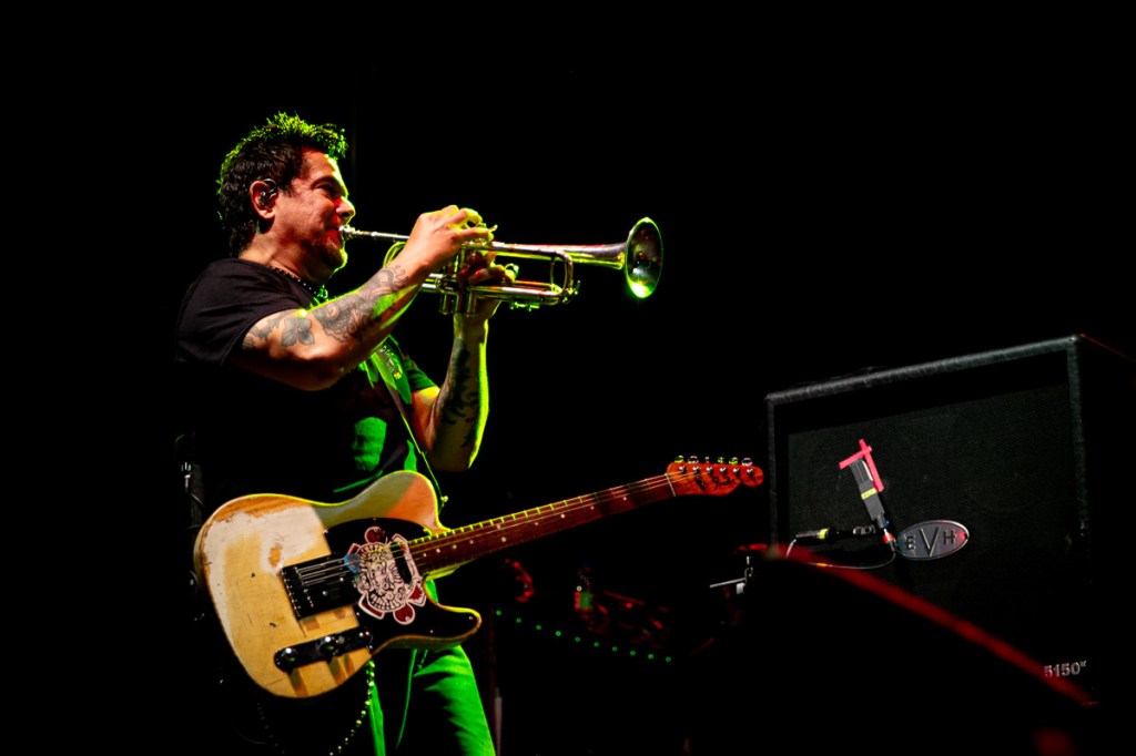 El Hefe plays trumpet while wearing a guitar.