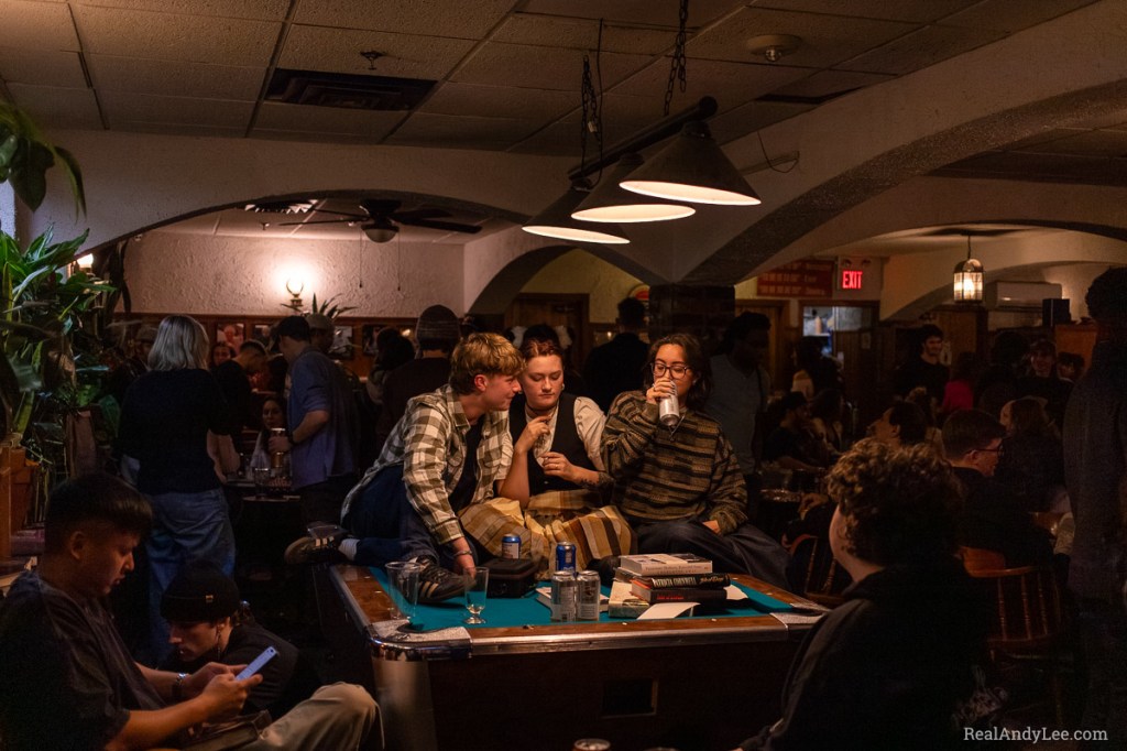 Three students sitting on a pool table with beer cans and books in a crowded bar