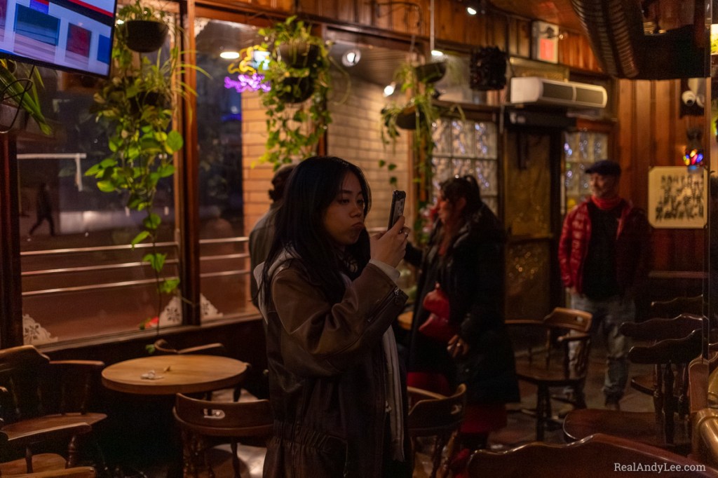 Asian woman wearing brown leather jacket pouting while taking a photo with her cell phone